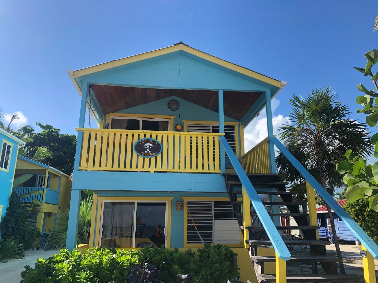 Exterior of a colorful blue and yellow cabana with stairs leading to second level, bright blue sky and tropical foliage