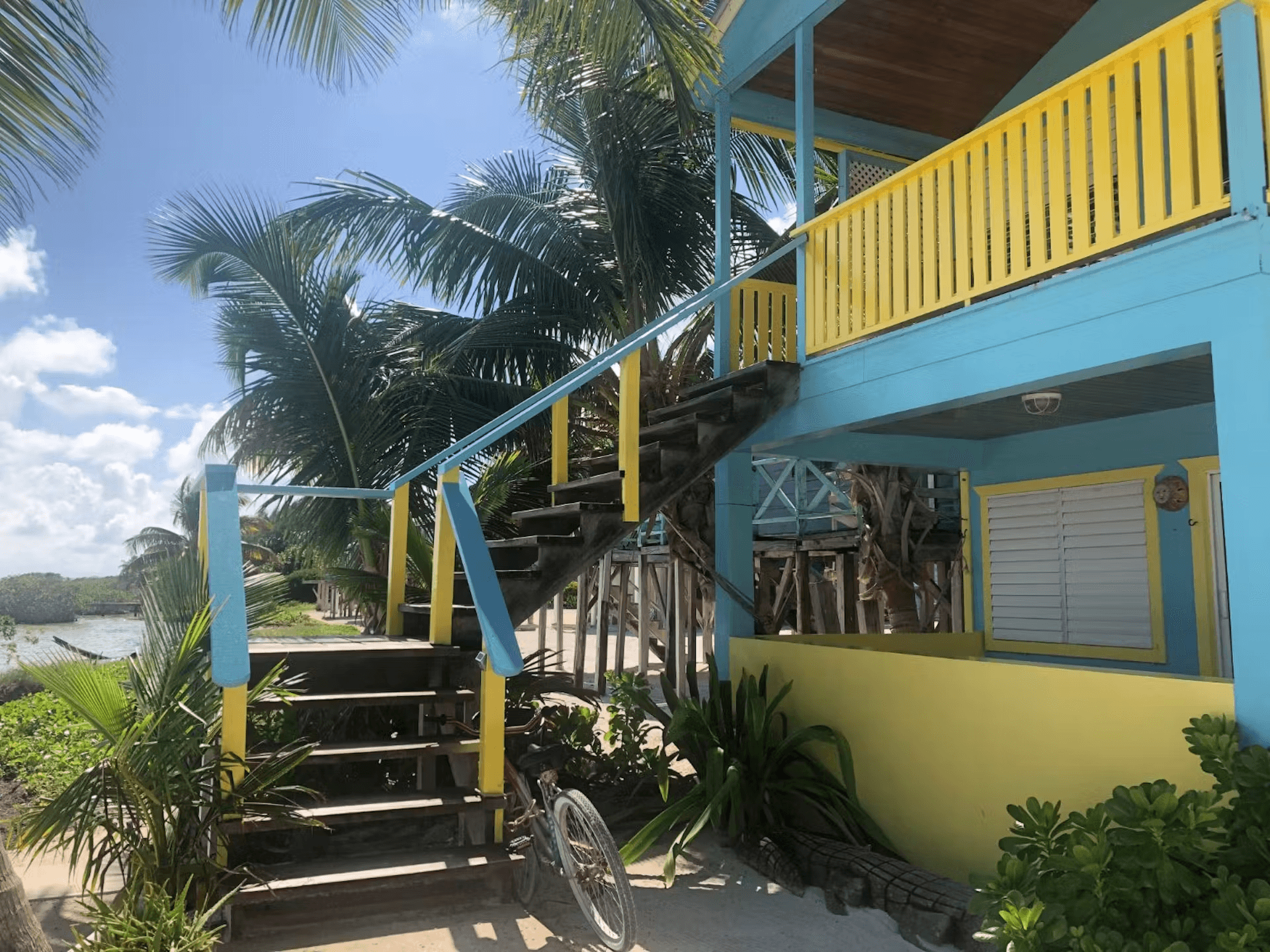 Exterior of a colorful blue and yellow cabana with stairs leading to second level, bright blue sky and tropical foliage