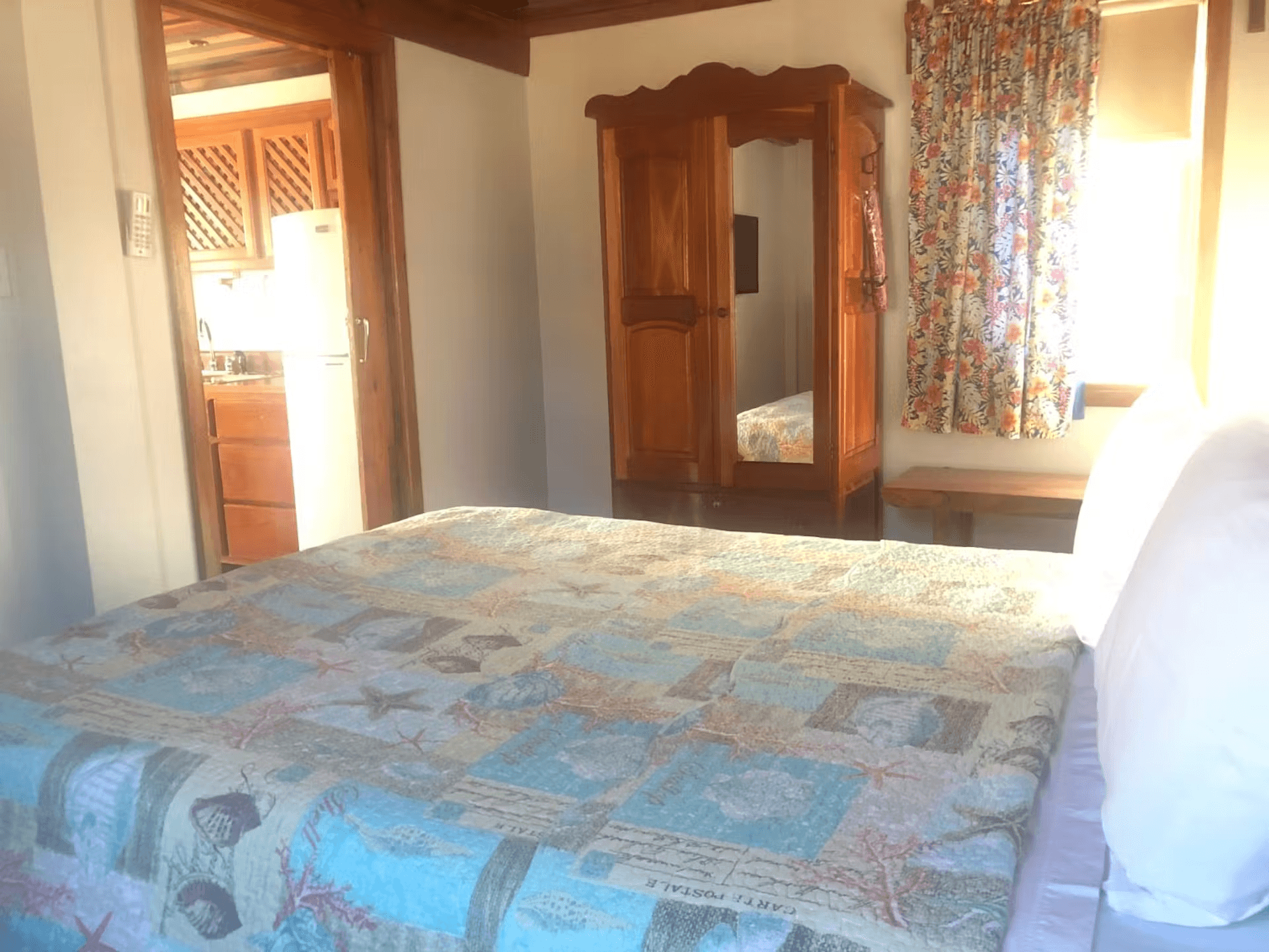 Bedroom with queen bed, wooden armoire, doorway leading into kitchen, and sunlight streaming through window