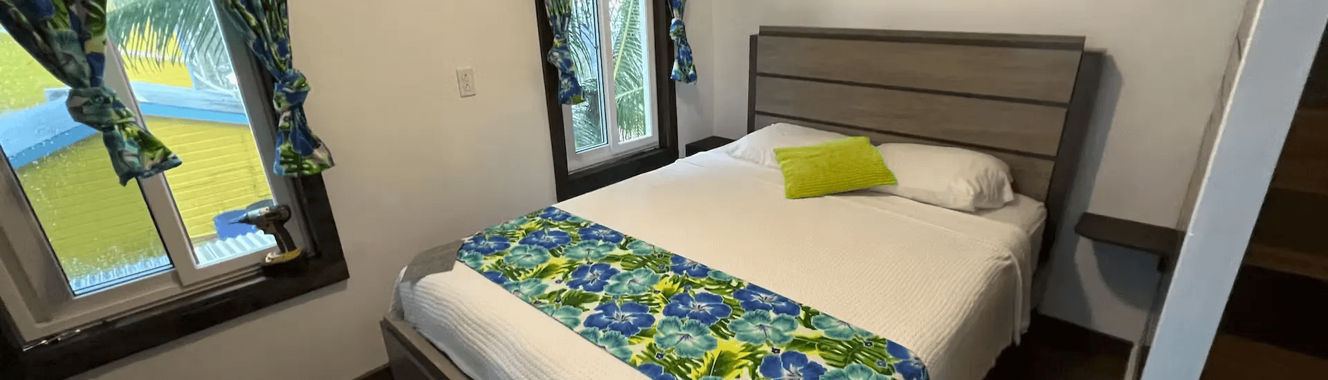 Gray wood slatted bed with white bed spread and a bright green and blue flowered throw across the foot of the bed, whight walls with tall windows and curtians to match the throw