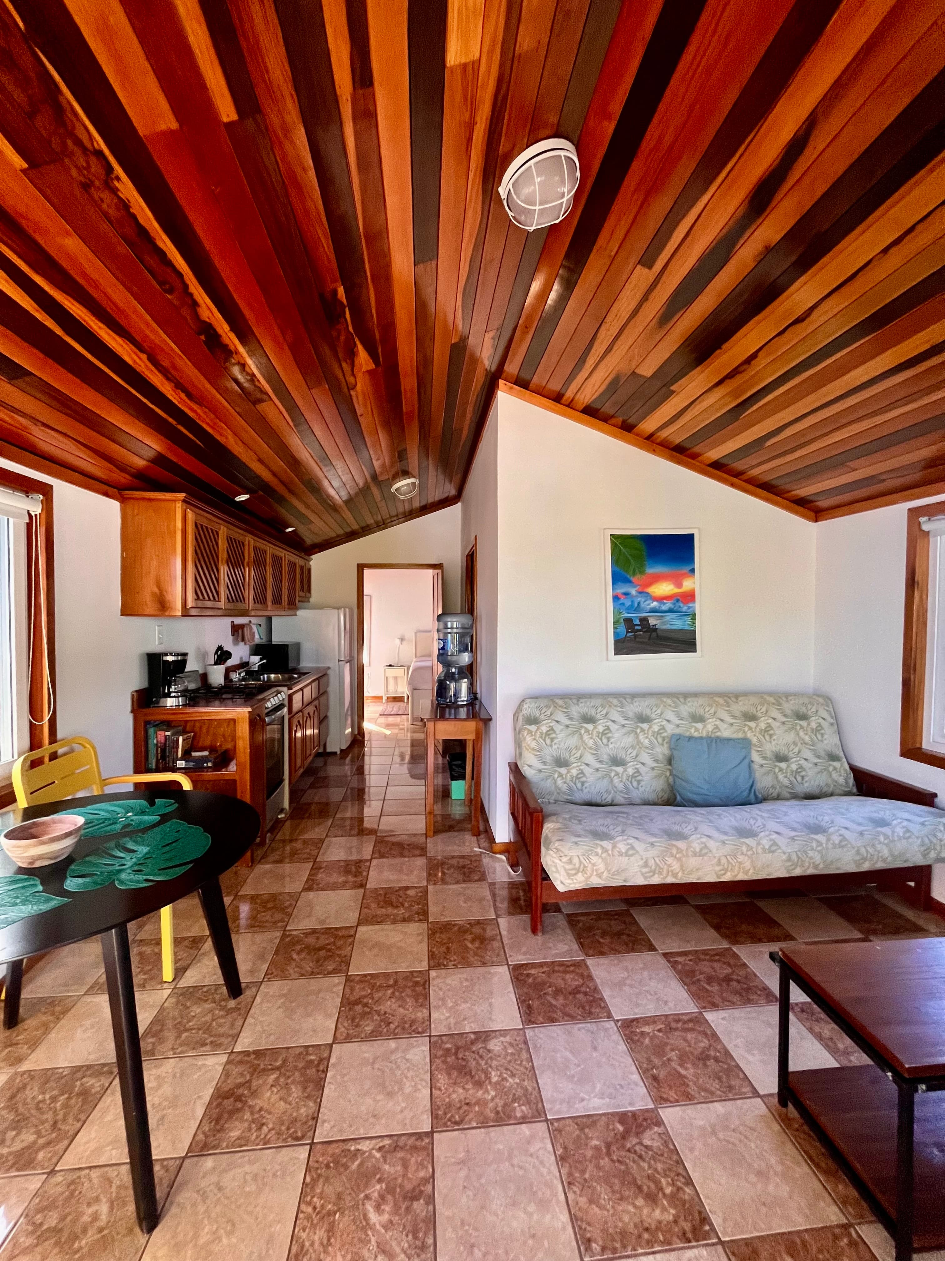 A cozy living space featuring a wooden ceiling, tiled floor, kitchen area, and a couch with decorative cushions.